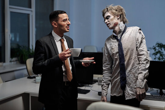 Young elegant businessman with cup of tea talking to male zombie in office
