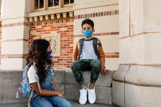 Young Students Wearing Masks Have A Conversation While Sitting On The Steps Their School