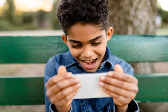 Boy Sitting on a Park Bench Reacts While Watching a Video on His Phone 