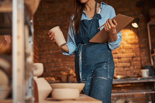 Young cheerful craft woman with clipboard in hands standing in art studio