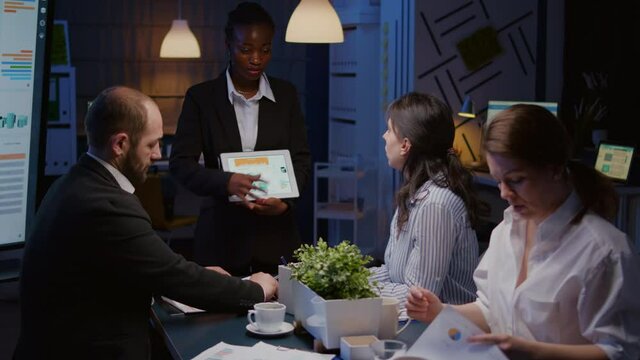 Entrepreneur woman with dark skin overworking in company office meeting room explaining management strategy using tablet. Workaholics multi-ethnic businesspeople brainstorming late at night