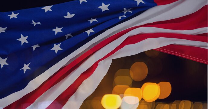 Composition of american flag billowing over defocussed bokeh orange and white lights on night sky