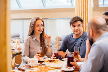 A group of young cheerful friends is sitting in a cafe talking and eating pizza. Lunch at the pizzeria.
