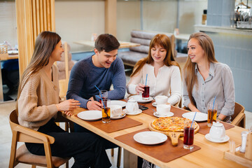 A group of young cheerful friends is sitting in a cafe talking and eating pizza. Lunch at the pizzeria.