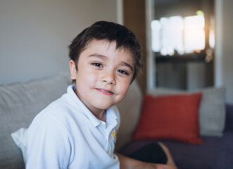 Closeup schoolboy looking at camera with smiling face, Happy Child relaxing at home after back from school, Indoor portrait positive kid sitting on sofa in living room