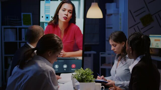 Workaholics overworked multi-ethnic businesspeople discussing with remote manager during online videocall conference meeting late at night in company office room. Focused teamwork working overtime