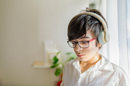 Close up pre-teen boy wearing white shirt, headphones, and glasses by a window