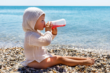 Cute caucasian little toddler child sits on the beach and enjoys drinking a drink children's mors at the sea on the beach in summer. © Татьяна Михайлина