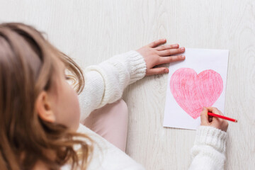 Little young girl draws a red heart on white paper on a wooden background floor. Top view. Copy space for text. Mother's Day. The child's love for his mother.