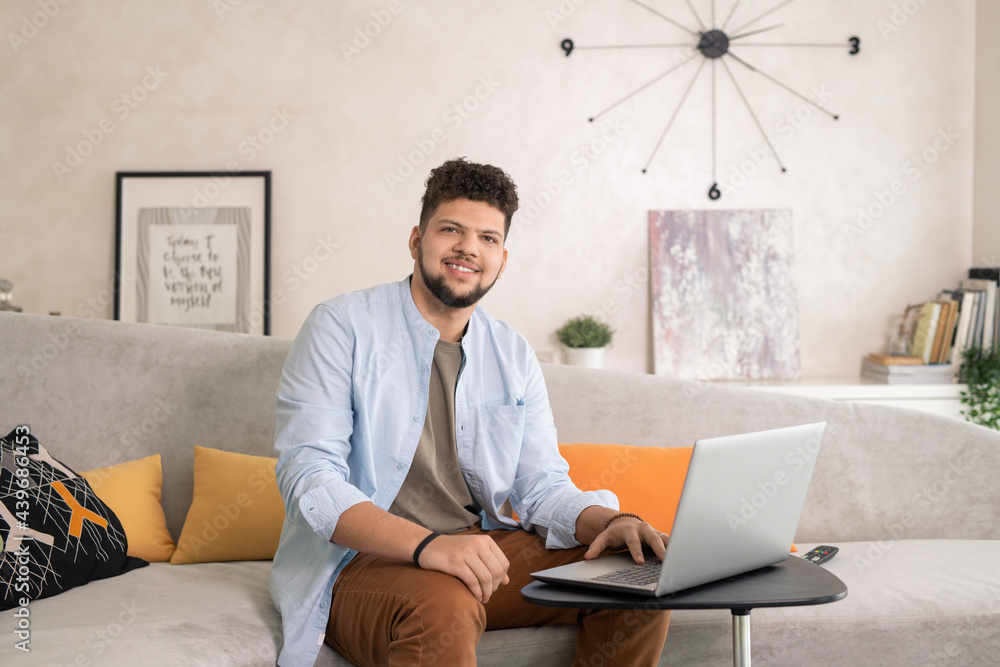 Poster young man in casualwear using laptop while working in home office