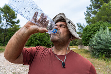 Caucasian hiker man wearing a hat and very sweaty, finishing water from a plastic bottle, on a very...