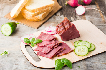 Fresh sliced marbled beef pastrami, cucumber, radish and basil on a cutting board. American delicacy. Rustic style