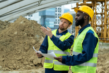 Two young intercultural builders in uniform discussing sketch and new construction