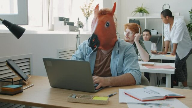 Funny man wearing horse mask is working with laptop typing concentrated on business activities in office. Crazy workplace and modern technology concept.