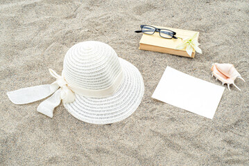 Summer still life. White straw hat on the sand of the mediterranean beach. Summer party invitation card mockup with white flower, shell, book and glasses. Vacation and relaxation concept.