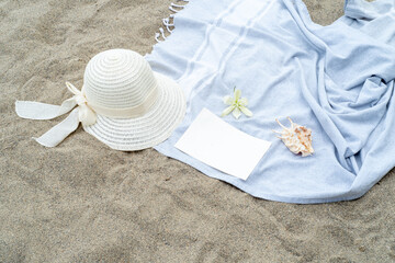 Fototapeta na wymiar Summer still life. White straw hat on the sand of the mediterranean beach. Summer party invitation card mockup with white flower, shell and blue towel. Vacation and relaxation concept.