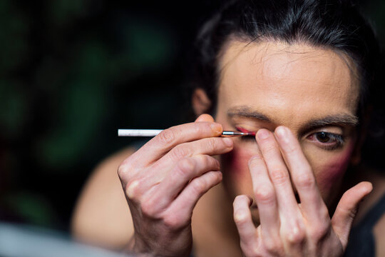 Young man looking in mirror and applying pink eyeshadow on eyelid while doing makeup in drag queen style