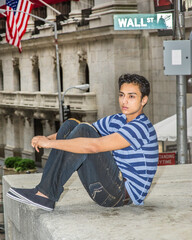 Dressing in a blue lines T shirt, a fashionable pants, and sitting on a stage by the street,  a young asian teenager is looking away and thinking.
