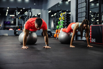 Fototapeta na wymiar Front view of a fitness couple in a plank position. Their feet are on a gray pilates ball while their hands are firmly touch on the black floor in an indore and spacious black gym or fitness studio