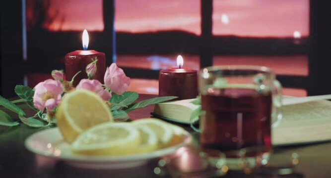 There are two burning candles on the table. Also on the table you can see: flowers, a plate with lemon, an open book and a glass of tea. Outside, you can see the river and the sunset.