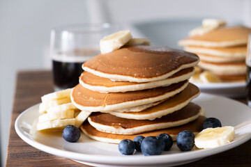 Stack of Tasty American style pancakes with a piece of butter, banana and blueberry on a white plate and a glass of black coffee . Breakfast for two or weekend brunch