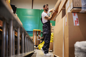 Hardworking tattooed bearded blue collar worker in overalls putting big heavy box on pile while standing in warehouse.