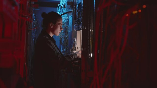 Side view of male Caucasian technician standing in blue and red-lighted data center room, typing on invisible computer keyboard