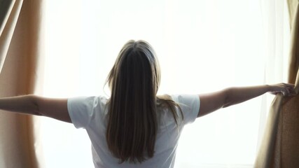 Happy optimistic young woman in white t-shirt, parting the beige curtains for the morning sun to illuminate her cozy apartment.She looks out the window alone and enjoys beautiful view of the cityscape