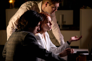 Side View On Confident Team Of Business People In Group, Checking Documents And Business Workflow Using pc Computer In New Startup Office At Night, Discussing, Looking At Screen Of Computer