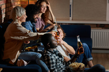 Multi-ethnic group of friends having small home party, meeting, sitting on couch, clinking bottles of beer, spending, enjoying time together, watching tv at night. side view