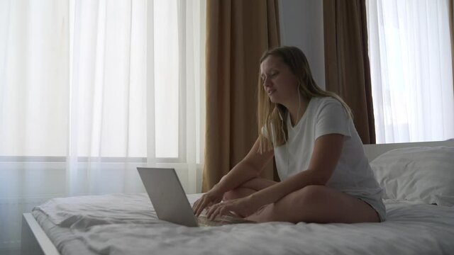 Woman in white T-shirt sits on the bed in headphones and works on notebook. Communicating in video conference on the computer, smiling and waving hand. The concept of work from home, quarantine