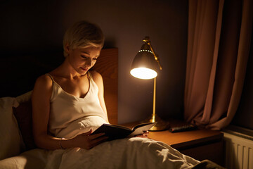 Beautiful female in pajamas lying on bed reading book, alone at night. Caucasian short haired lady in bedroom, charming cute woman in room lighted by lamp - obrazy, fototapety, plakaty