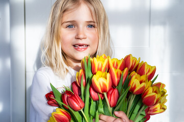 smiling toothless little girl with a huge bunch of tulips on the white background with straight sunlight and shadows. Getting flowers. Gift bouquet for Mother's day, Birthday surprise. Happy childhood
