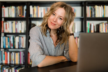 Woman Smiles at the Camera While Working on Her Laptop in Front of a Bookshelf 