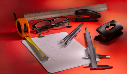 various measurement and design elements on a red table (gauge, meter, ruler, button, stapler, glasses, hole punch, pencil and pen)