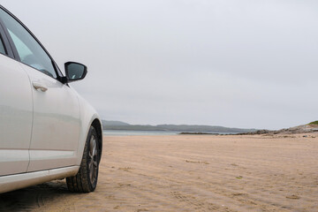 Fototapeta na wymiar White car parked on a sandy beach by the ocean. Omey island, county Galway, Ireland, cloudy sky, Nobody, Travel in a rented car concept. Copy space