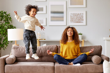 Young african woman mother in lotus pose meditating while child playing on background in living room