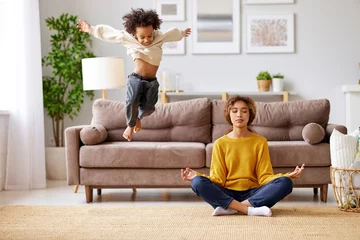  Young african woman mother in lotus pose meditating while child playing on background in living room © JenkoAtaman
