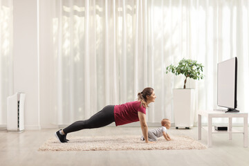 Mother doing plank exercises with her baby crawling on carpet in front of tv