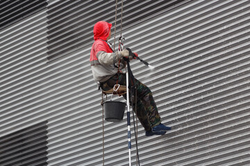 Climber on cables with a pressure washer cleans the facade of a gray building.