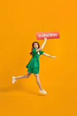 Female Blogger asks to subscribe to channel for video vlogs. Redhead lady in green dress emotionally smiling at camera, jumping, going or walking, hurry. isolated on yellow background in studio