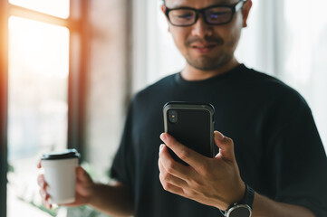 Happy Asian man using smartphone and drinking coffee he's smiling and relax in living room at home, man using cellphone for searching data and social media on internet. Technology and people concept.