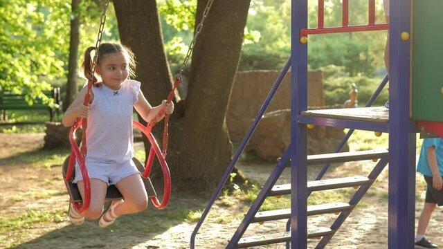 Close-up. A little beautiful girl with ponytails on her head swings on a swing on a bright sunny day in the park. Children's games. Children's playground. Children's complex for fun. Slow Motion