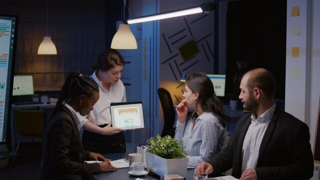 Workaholics diverse multi-ethnic businesspeople analyzing financial graphs using tablet computer overworking in company meeting room. Overworked manager brainstorming ideas in evening