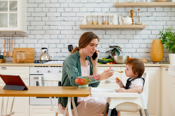 Focused mother feeding toddler son and talking via smartphone