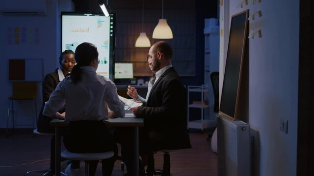 Selective focus of diverse multi-ethnic businesspeople overworking late at night analyzing management ideas solving company statistics. Workaholics teamwork working in office meeting room in evening