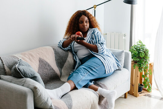 Woman on the couch eating apple holding tv remote
