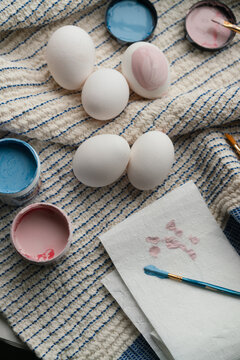 Five eggs on  striped tea towel with paint brushes and pink and blue colours  next to them 
