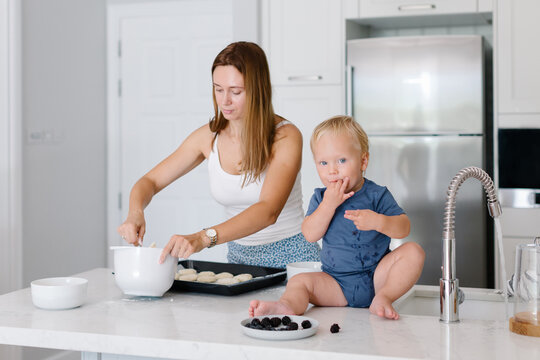 Mother cooking and her toddler is sitting next to her on a countertop and eating berries