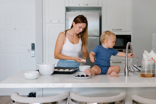 Mother cooking and her toddler is sitting next to her on a countertop 